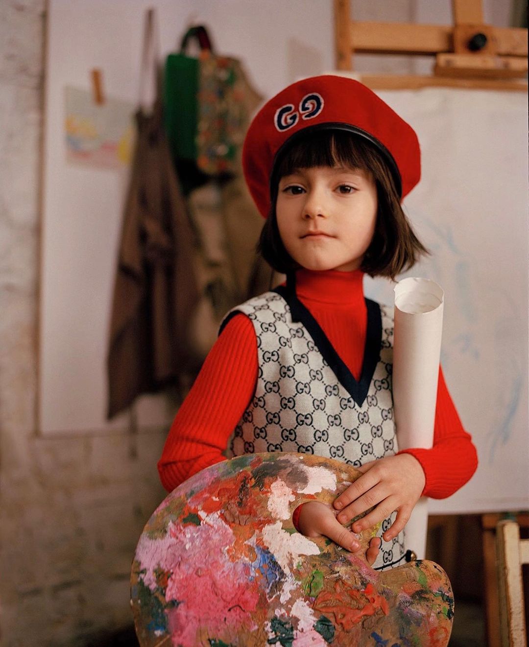 Child artist with paint palette, beret, and Gucci vest in front of a canvas in an art studio setting.