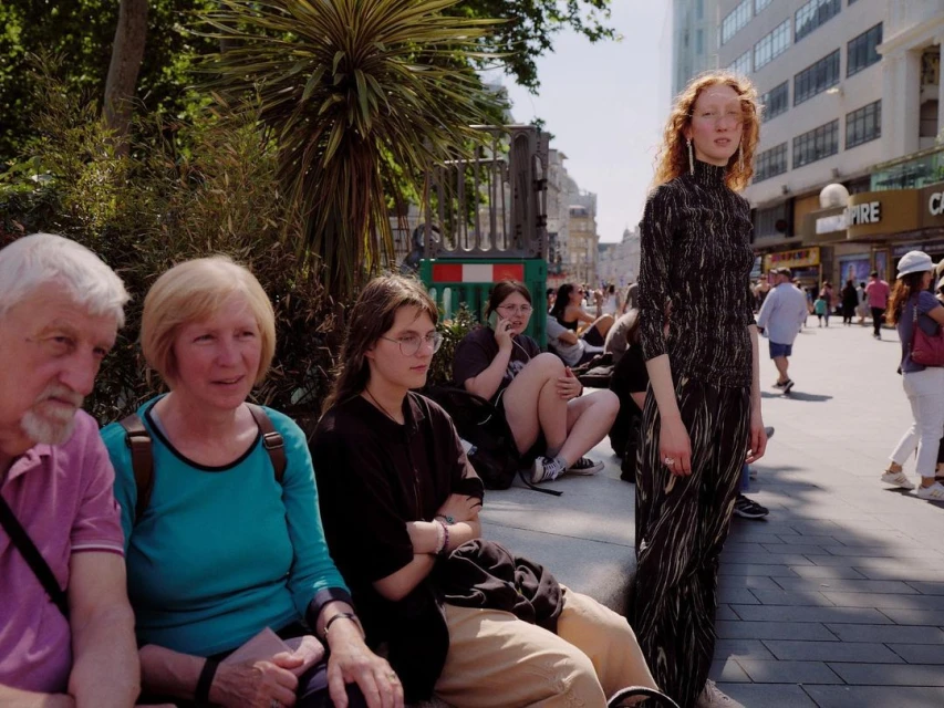Outdoor shot of red-haired woman in black dress standing near seated group of people in casual clothing on a sunny urban day.