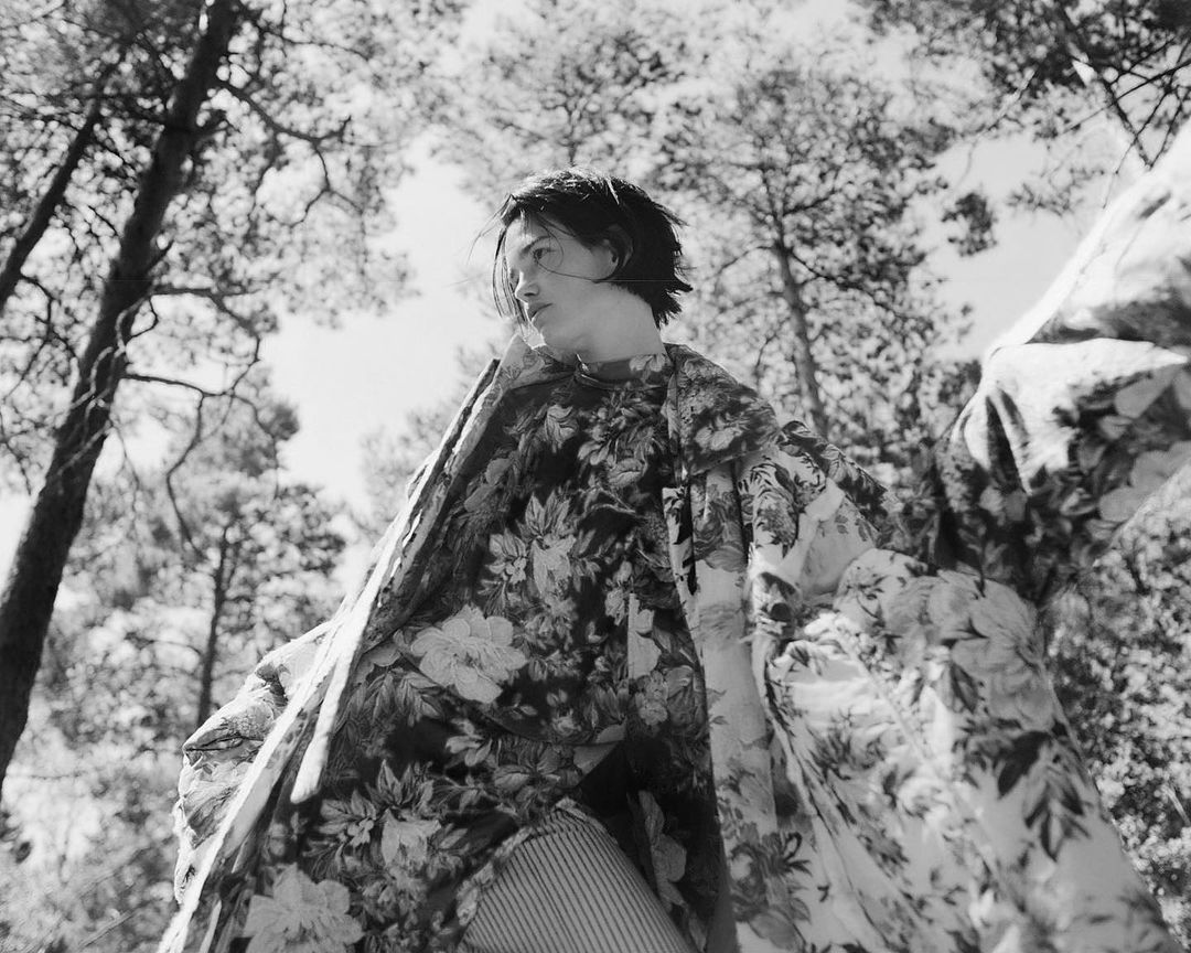 Woman wearing a floral print jacket in an outdoor forest setting, captured in a black and white photograph with a low-angle perspective.
