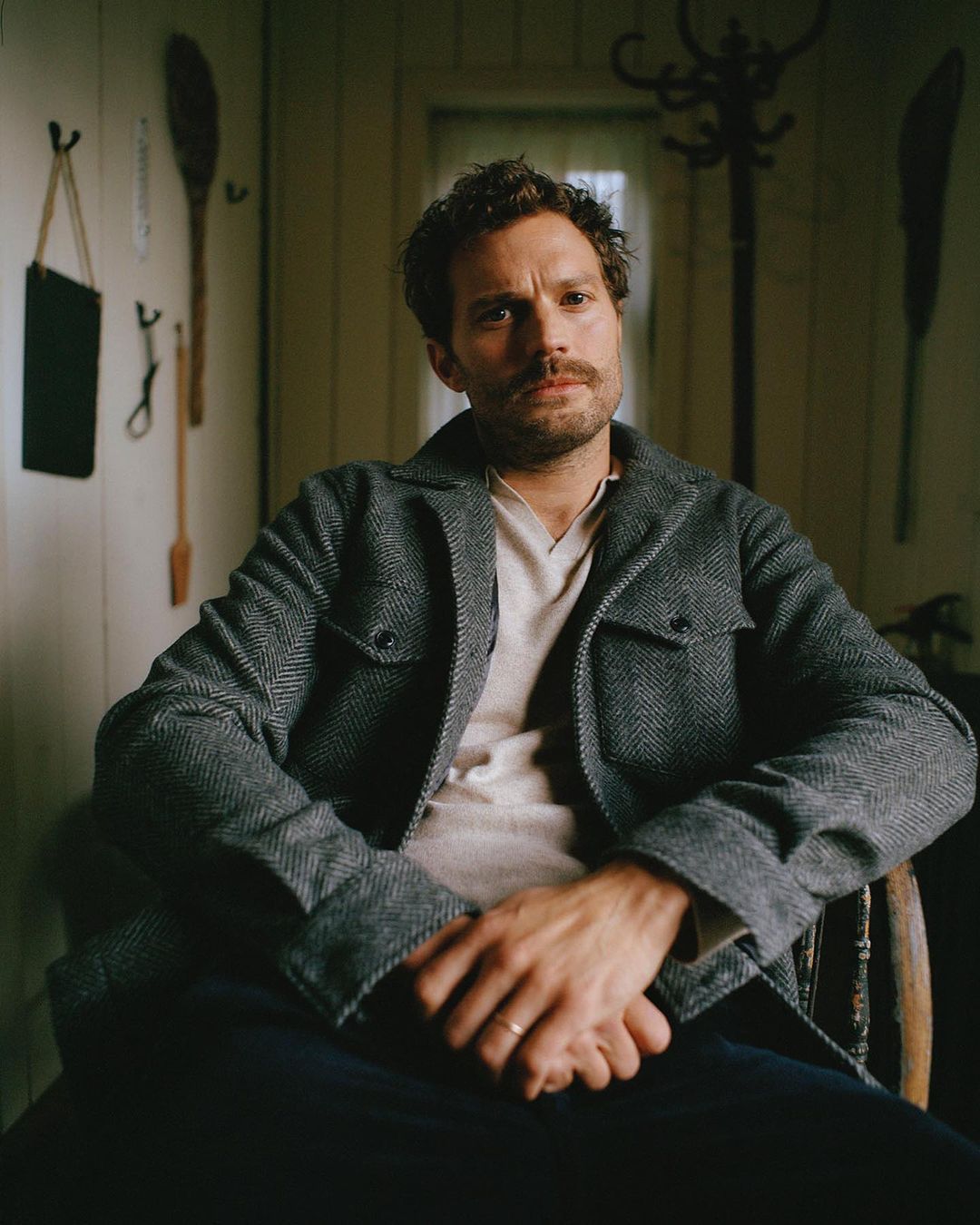 Portrait of a fair-skinned adult male with dark hair sitting in a wood chair wearing a gray wool coat inside a rustic room.