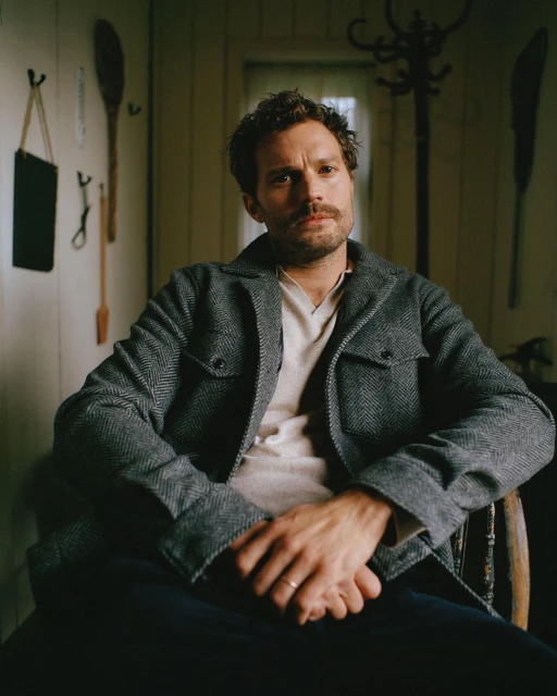 Portrait of a fair-skinned adult male with dark hair sitting in a wood chair wearing a gray wool coat inside a rustic room.