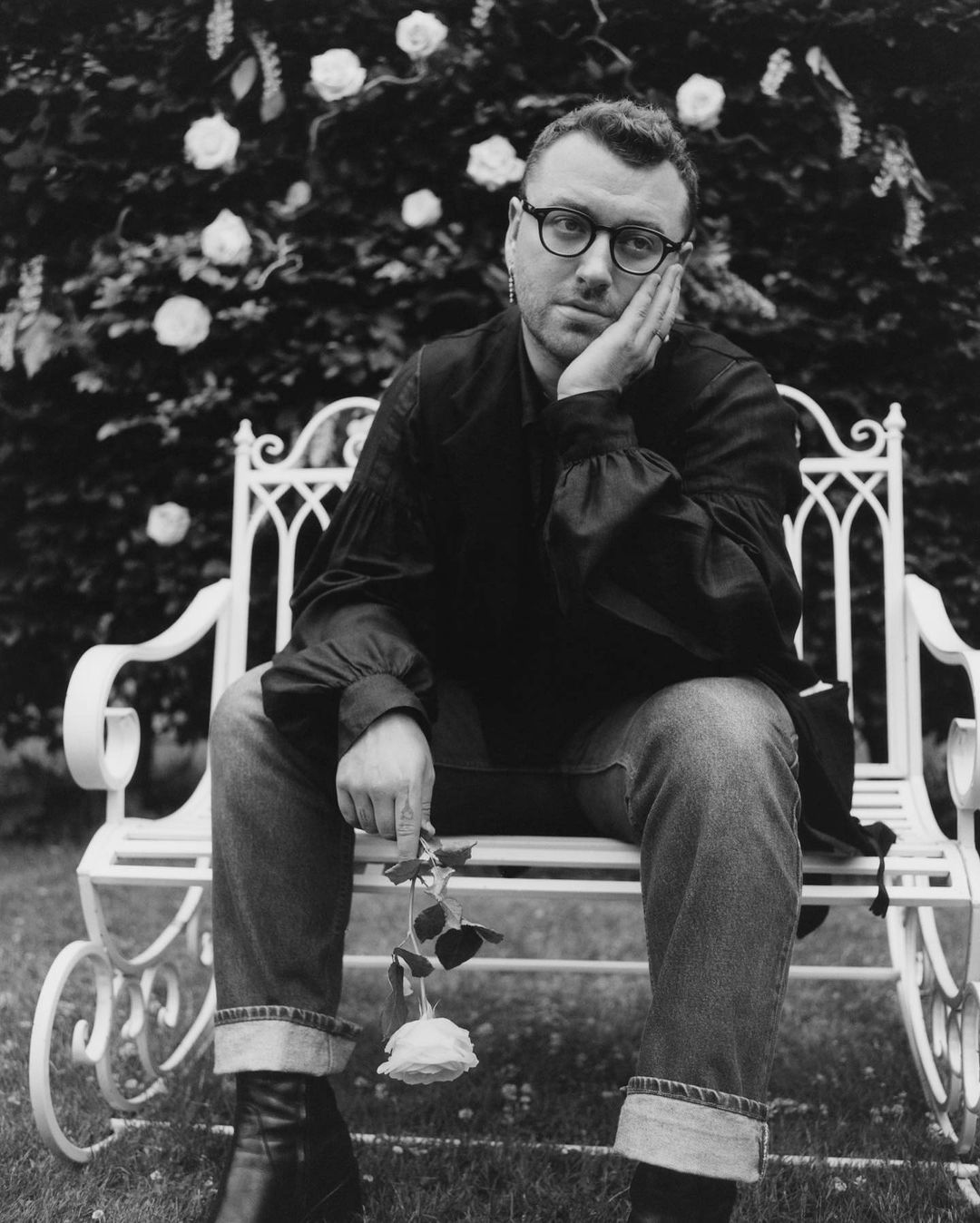 Black and white portrait of a man with glasses sitting on a bench holding a rose, with a rose bush as the backdrop.