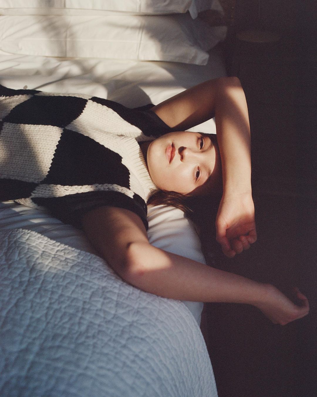 Woman lying on a white bed in a checkerboard sweater, resting with her arm raised and her gaze towards the camera in soft lighting.
