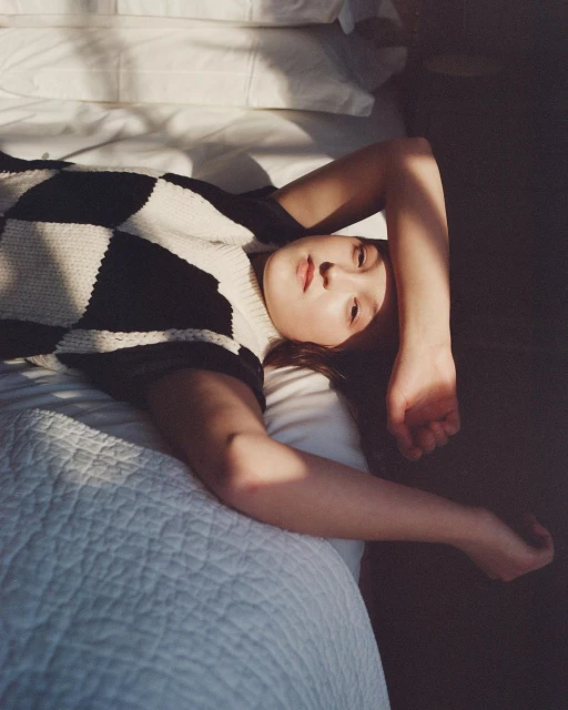 Woman lying on a white bed in a checkerboard sweater, resting with her arm raised and her gaze towards the camera in soft lighting.