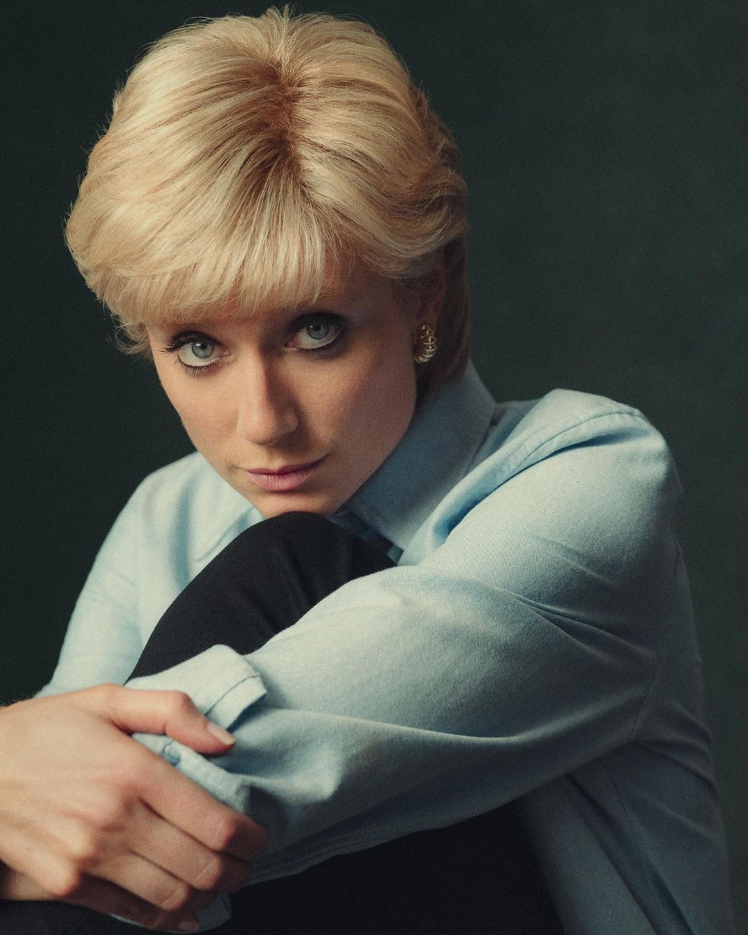 Portrait of an adult woman with blond hair, wearing a light blue shirt, embracing her knees indoors with a dark background.