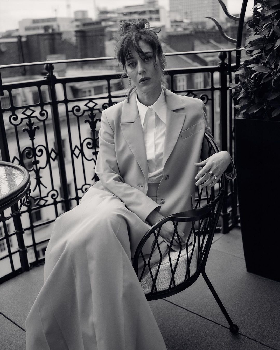 Monochrome portrait of a woman in a suit sitting on a chair on a balcony with an urban background, captured in soft lighting.
