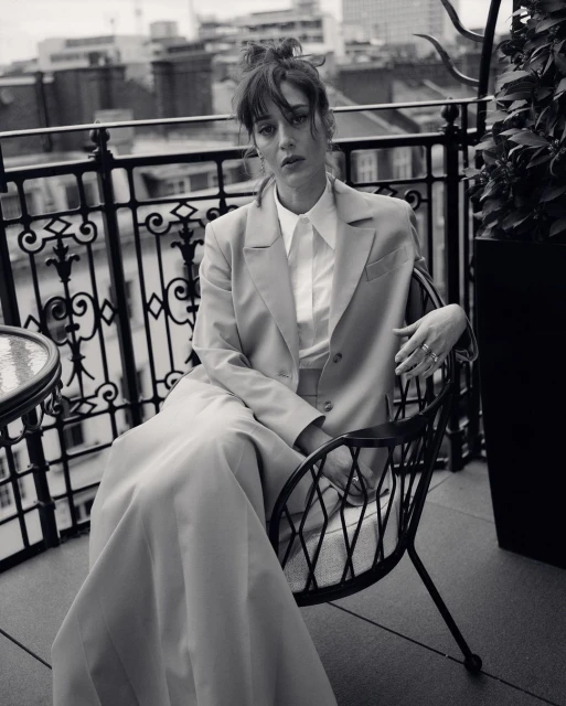Monochrome portrait of a woman in a suit sitting on a chair on a balcony with an urban background, captured in soft lighting.