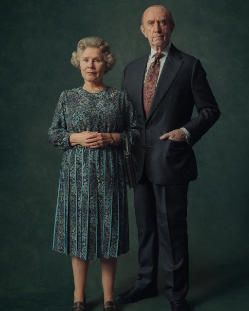 Portrait of two senior adult actors wearing a suit and patterned dress, posing in front of an dark green studio backdrop for a formal photo.