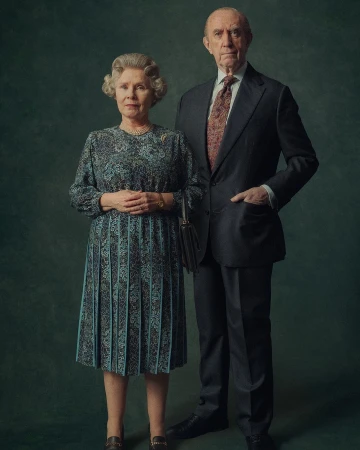 Portrait of two senior adult actors wearing a suit and patterned dress, posing in front of an dark green studio backdrop for a formal photo.