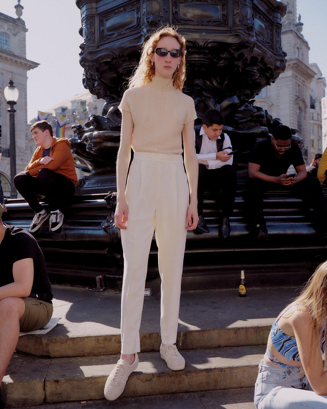 A tall, slender person wearing sunglasses stands in an urban square in neutral attire. The model is near a large fountain.