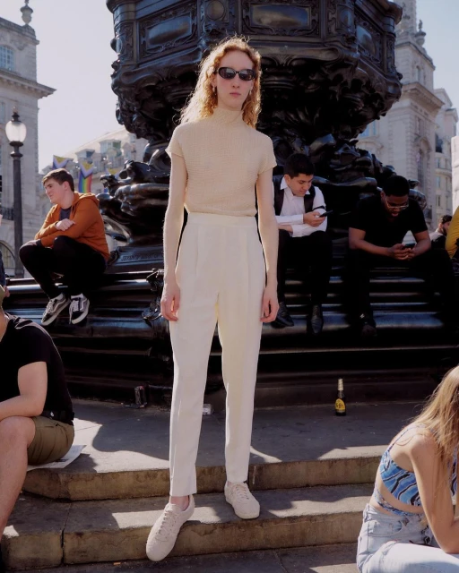 A tall, slender person wearing sunglasses stands in an urban square in neutral attire. The model is near a large fountain.