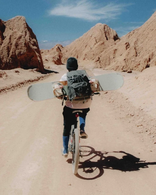 Adult rides bicycle with snowboard attached to backpack through desert canyon under blue sky on sunny day.
