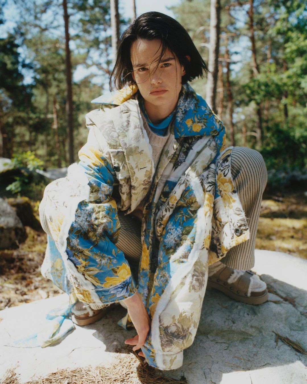 Young adult crouches on rock in forest wearing floral print jacket and striped pants, glancing at the camera with a neutral expression.