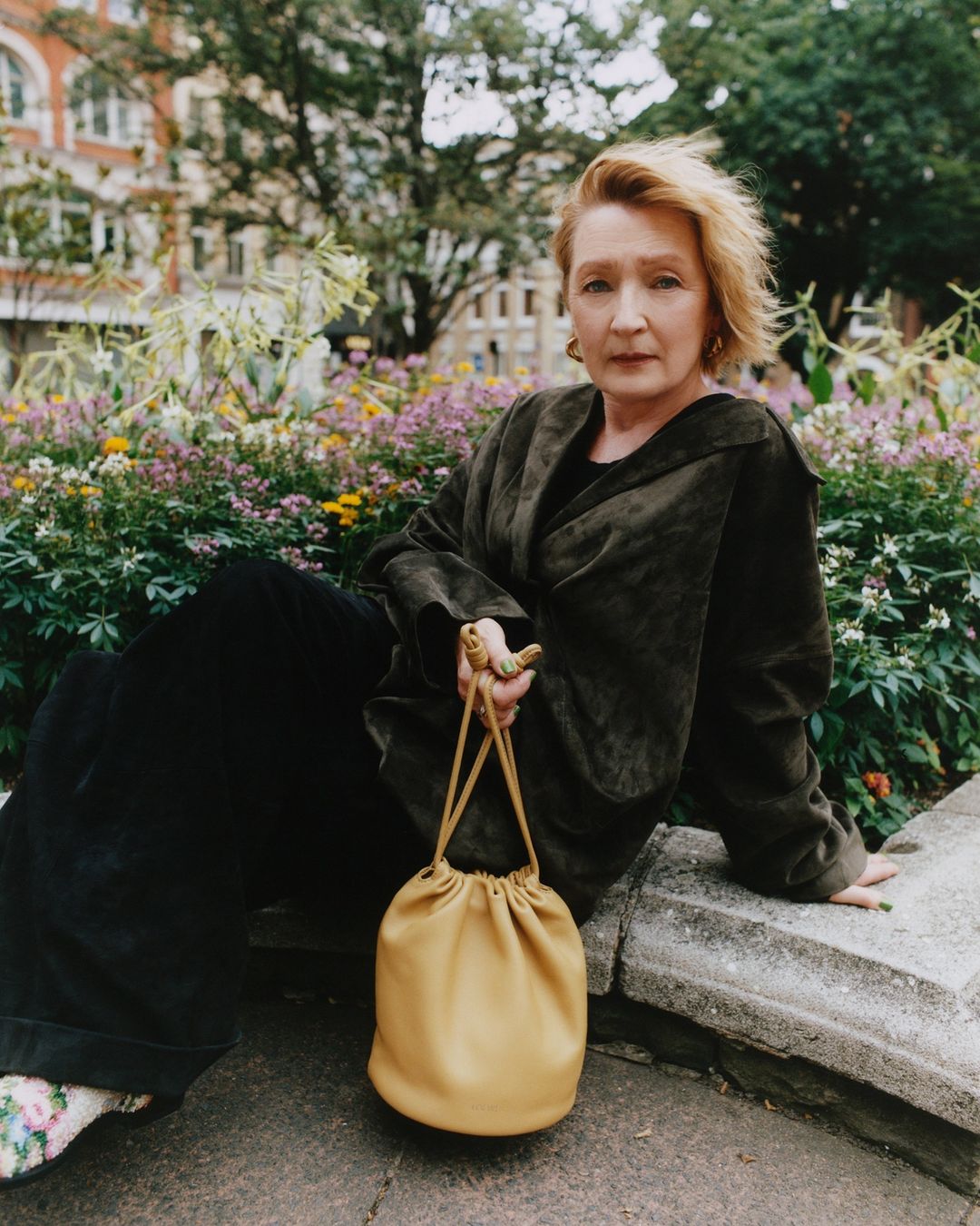 Mature woman with short blonde hair sits outside wearing a green jacket with black pants and a tan purse in a park setting.