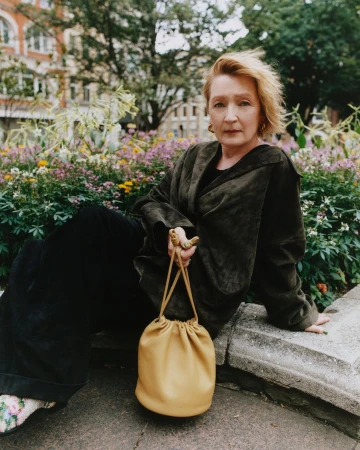 Mature woman with short blonde hair sits outside wearing a green jacket with black pants and a tan purse in a park setting.