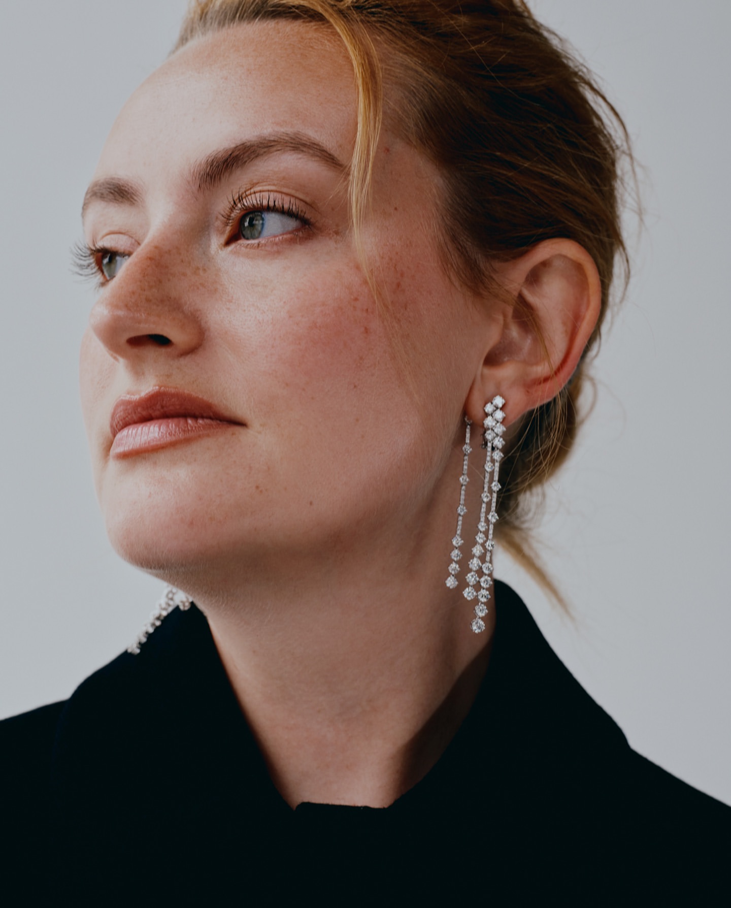 Close-up of a woman with fair skin and light brown hair wearing diamond drop earrings and a black jacket against a neutral background.