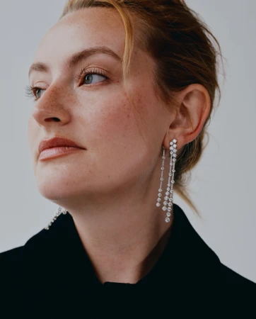Close-up of a woman with fair skin and light brown hair wearing diamond drop earrings and a black jacket against a neutral background.
