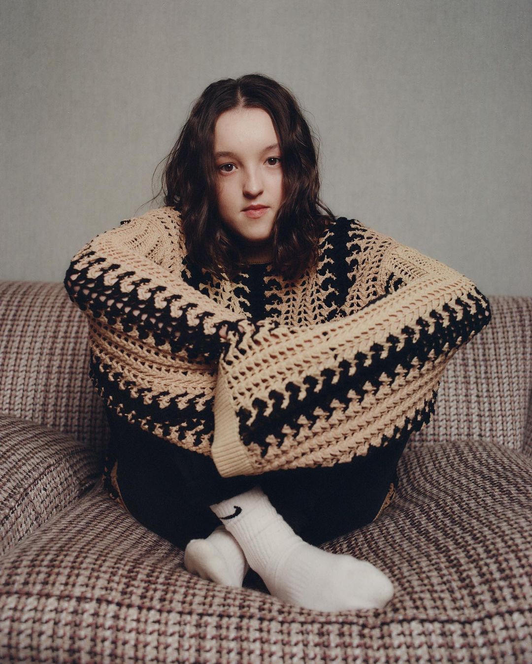 Portrait of a young woman with curly brunette hair sitting on a patterned couch, wearing a knit sweater and socks with a logo.