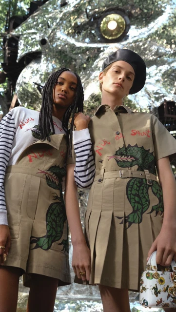 Two women pose in coordinated khaki outfits with green dragon designs in an outdoor setting, with a mirrored structure behind them.