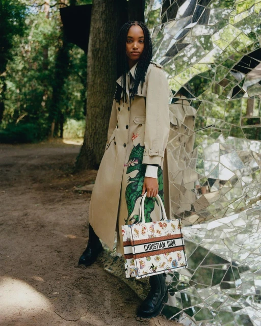 A young Black woman stands next to a modern sculpture, showcasing a beige trench coat with dragon print and a Christian Dior tote bag in a forest.