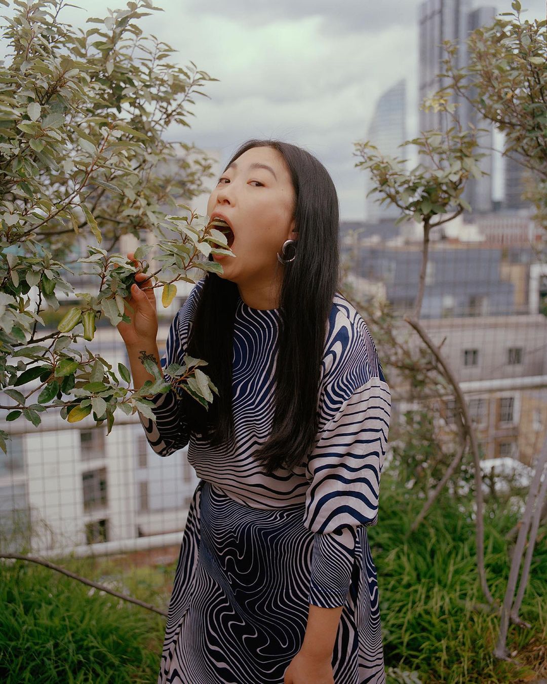 Asian woman in patterned dress eating leaves outdoors on a rooftop garden in a high-angle medium shot, city buildings in background.