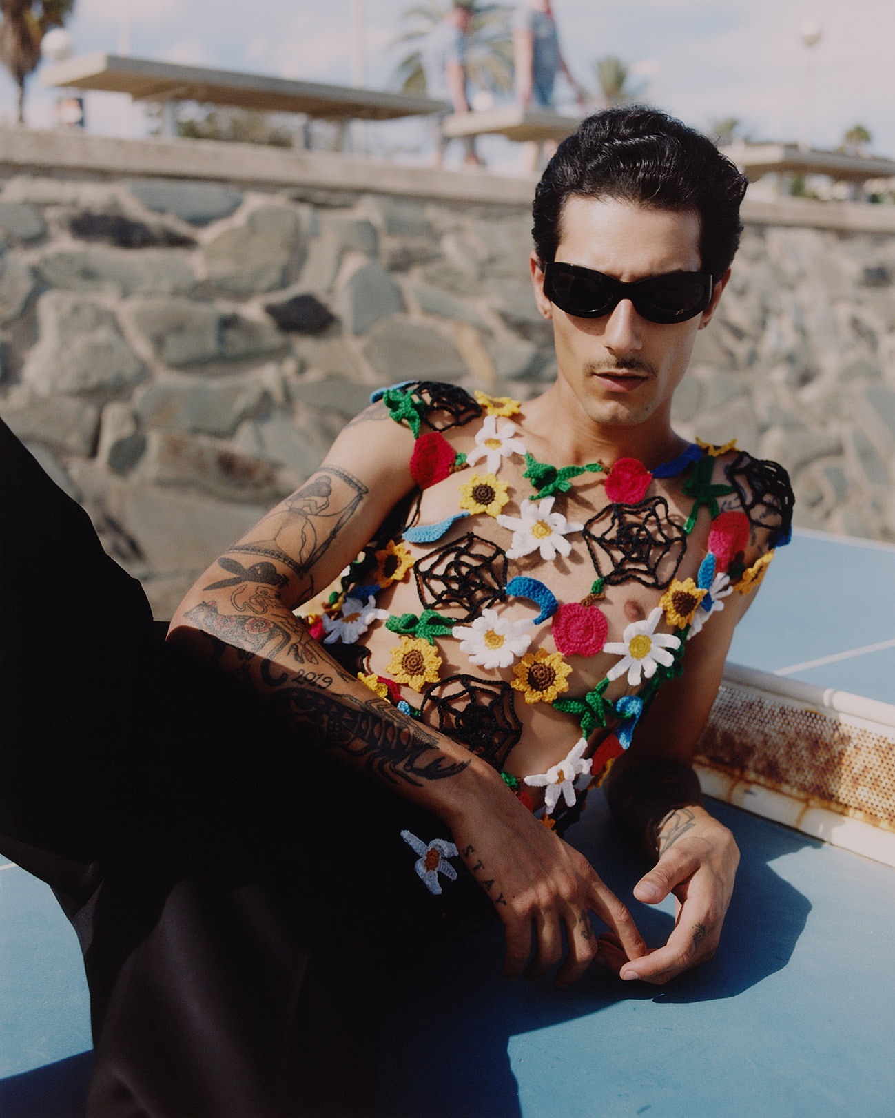 Adult male poses with sunglasses and a floral crochet top against a textured wall near a ping pong table outside in daylight.