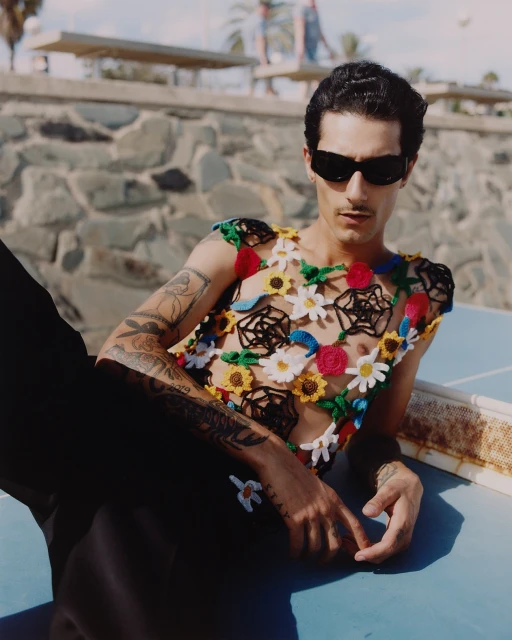 Adult male poses with sunglasses and a floral crochet top against a textured wall near a ping pong table outside in daylight.