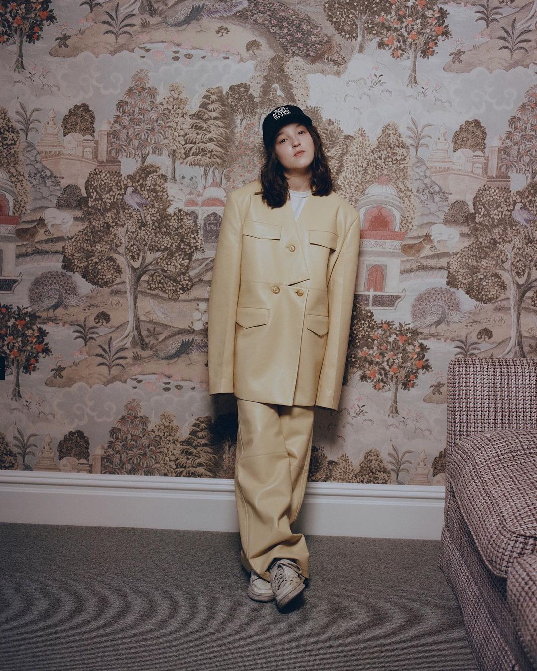 Woman in light yellow suit and baseball cap poses against wallpaper with landscape imagery in a medium, eye-level indoor shot.