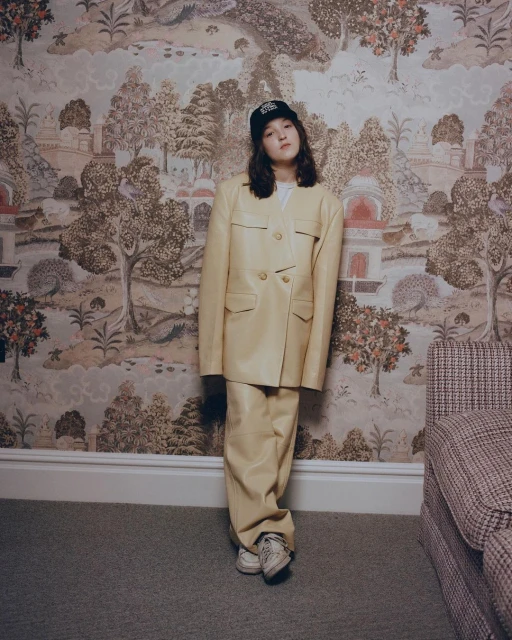 Woman in light yellow suit and baseball cap poses against wallpaper with landscape imagery in a medium, eye-level indoor shot.