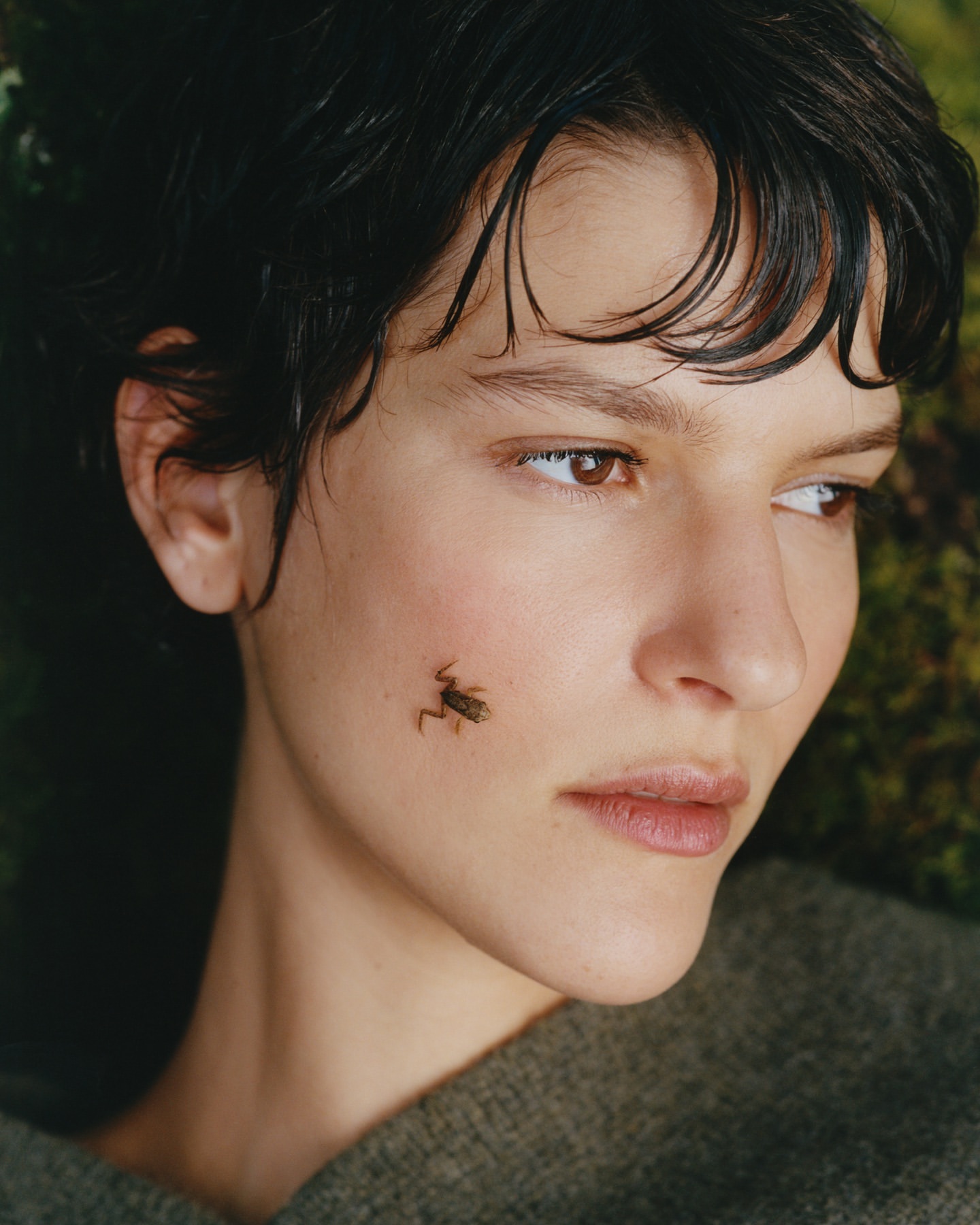 Close-up portrait of a woman with a short dark hairstyle and a grasshopper resting on her cheek against a background of green moss.