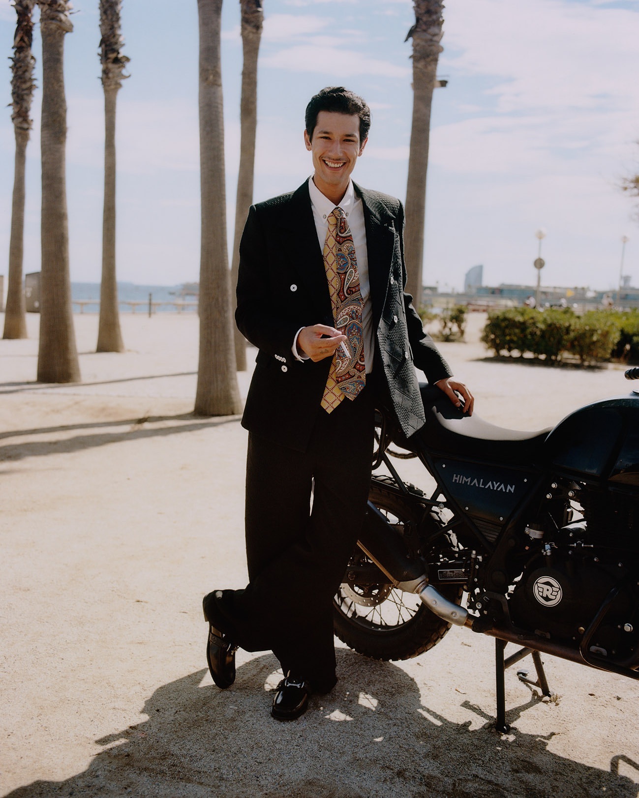 Smiling young man in a black suit and patterned tie leaning against a motorcycle on a sandy path with palm trees