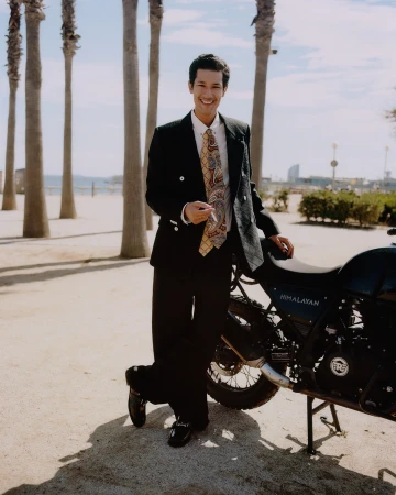 Smiling young man in a black suit and patterned tie leaning against a motorcycle on a sandy path with palm trees