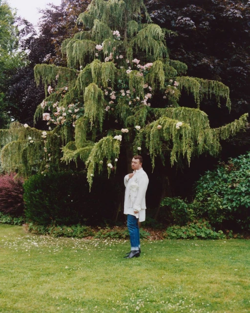 Adult stands in green lawn next to a weeping evergreen with light pink flowers, wearing jeans and a white overcoat in daylight.