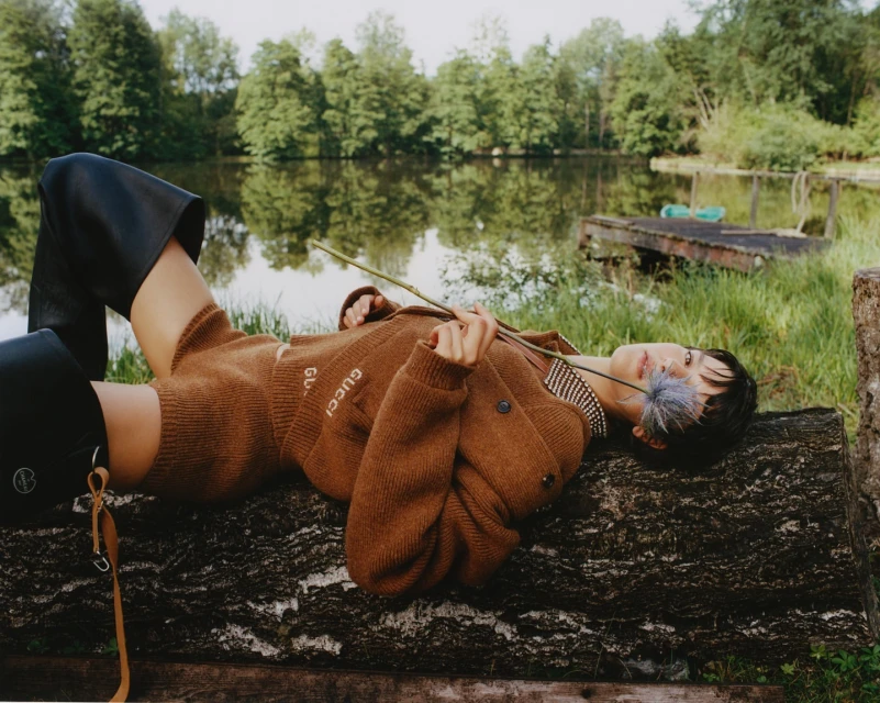 A woman lies on a log by a lake wearing a brown Gucci cardigan and black boots, holding a blue flower, in an outdoor fashion editorial.