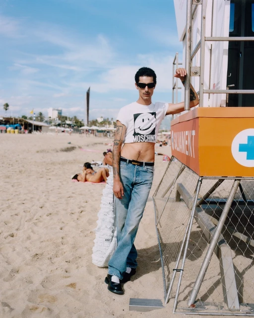 A slender man in a crop top stands next to an orange lifeguard tower on a sunny beach, with blurred people and buildings in background.
