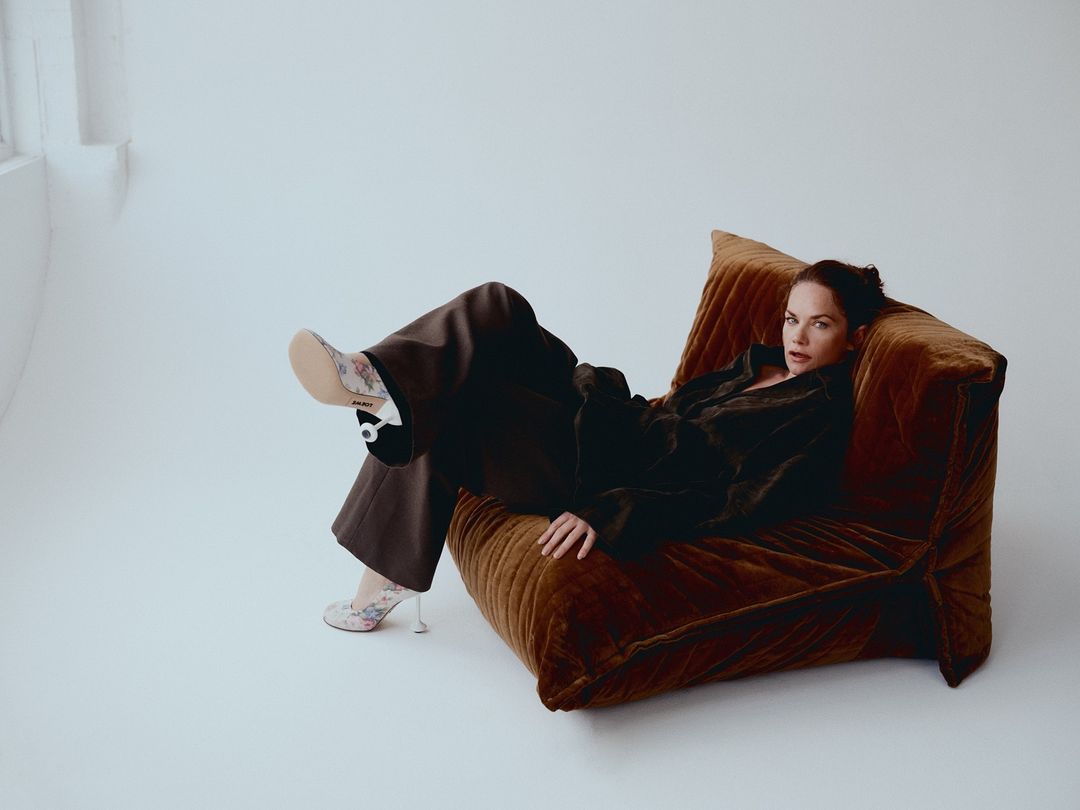 Woman in velvet jacket and brown pants reclines on brown velvet armchair in a photo studio with natural light from the left.