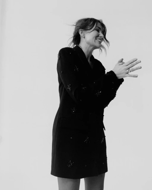 Monochrome portrait of a smiling woman with brown hair, wearing a beaded black blazer dress in front of a plain white backdrop.