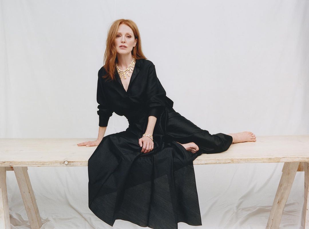 Julianne Moore seated on a wooden trestle table wearing a black dress and gold statement necklace against a white background.