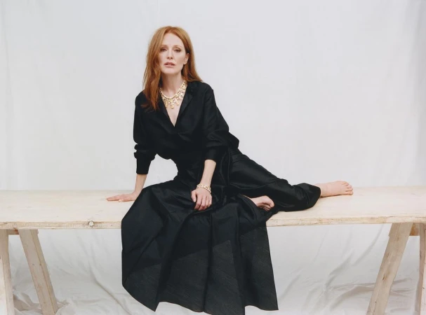 Julianne Moore seated on a wooden trestle table wearing a black dress and gold statement necklace against a white background.