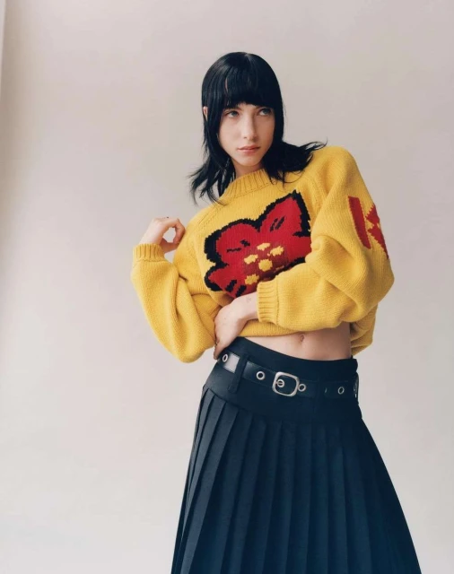 A stylish young woman poses indoors wearing a yellow sweater with a large floral design and a black pleated skirt with a belt.