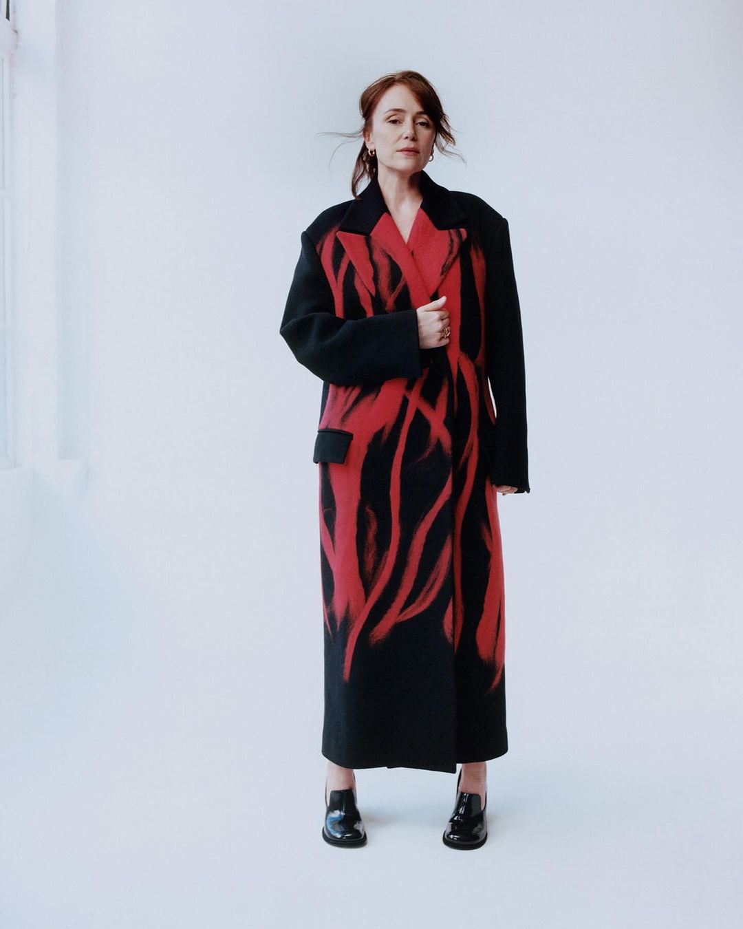 Adult woman poses in a floor-length coat with a red graphic design, complemented by black shoes in a bright photo studio.