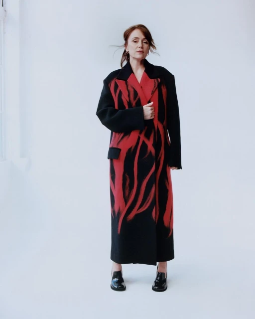 Adult woman poses in a floor-length coat with a red graphic design, complemented by black shoes in a bright photo studio.