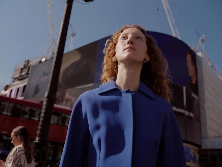 A woman in a blue coat looks up in an urban setting, with a blurred bus and digital billboards in the background under a clear blue sky.