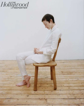 A person with short dark hair wearing a white shirt and trousers sitting on a wooden chair against a plain wall.