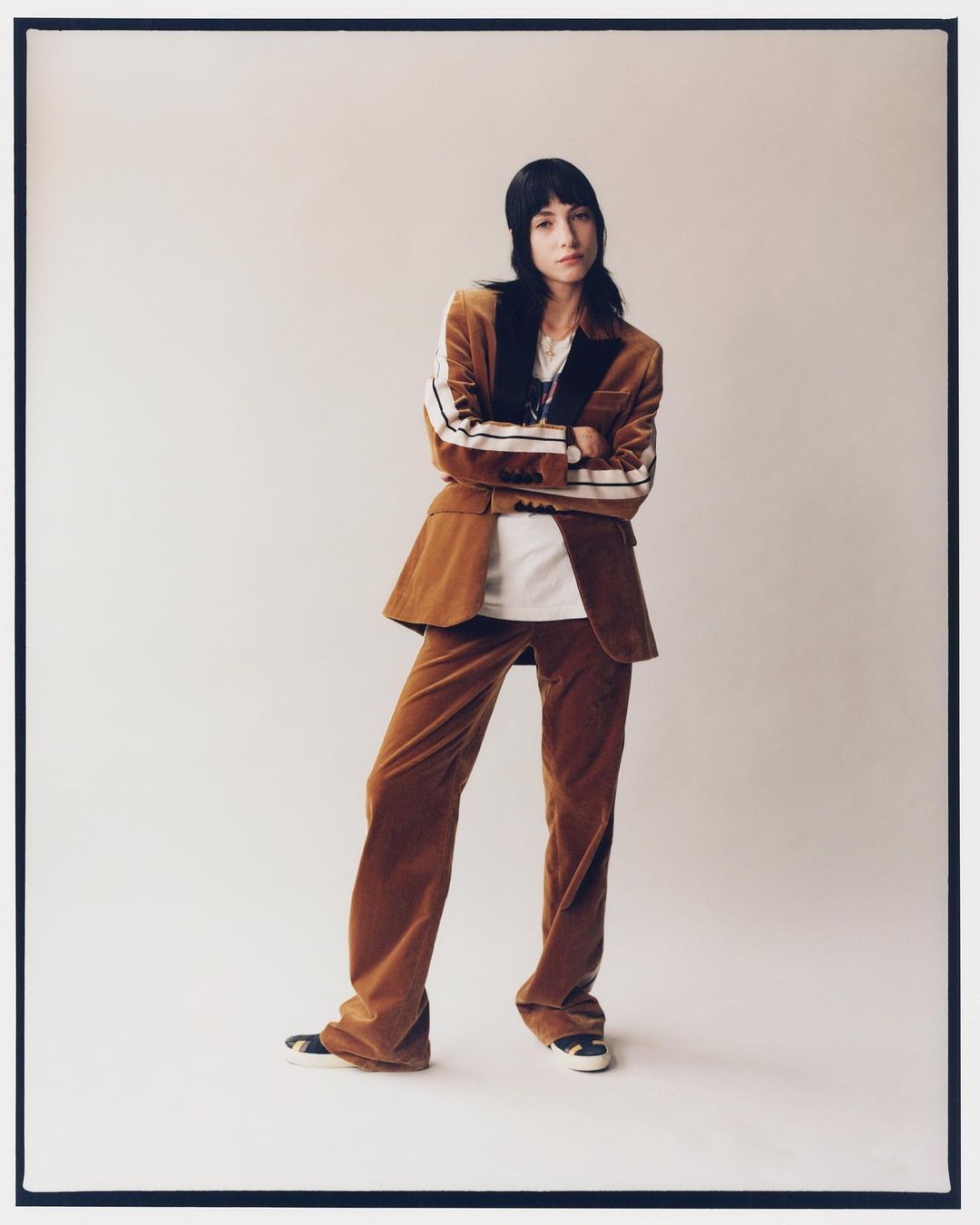 A woman in a brown corduroy suit stands in a photo studio, arms crossed, gazing at the camera in a full-length fashion shot.