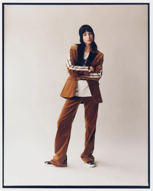 A woman in a brown corduroy suit stands in a photo studio, arms crossed, gazing at the camera in a full-length fashion shot.