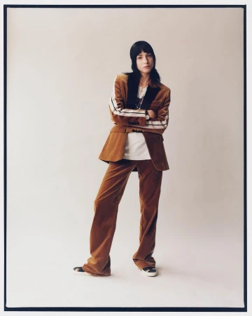 A woman in a brown corduroy suit stands in a photo studio, arms crossed, gazing at the camera in a full-length fashion shot.
