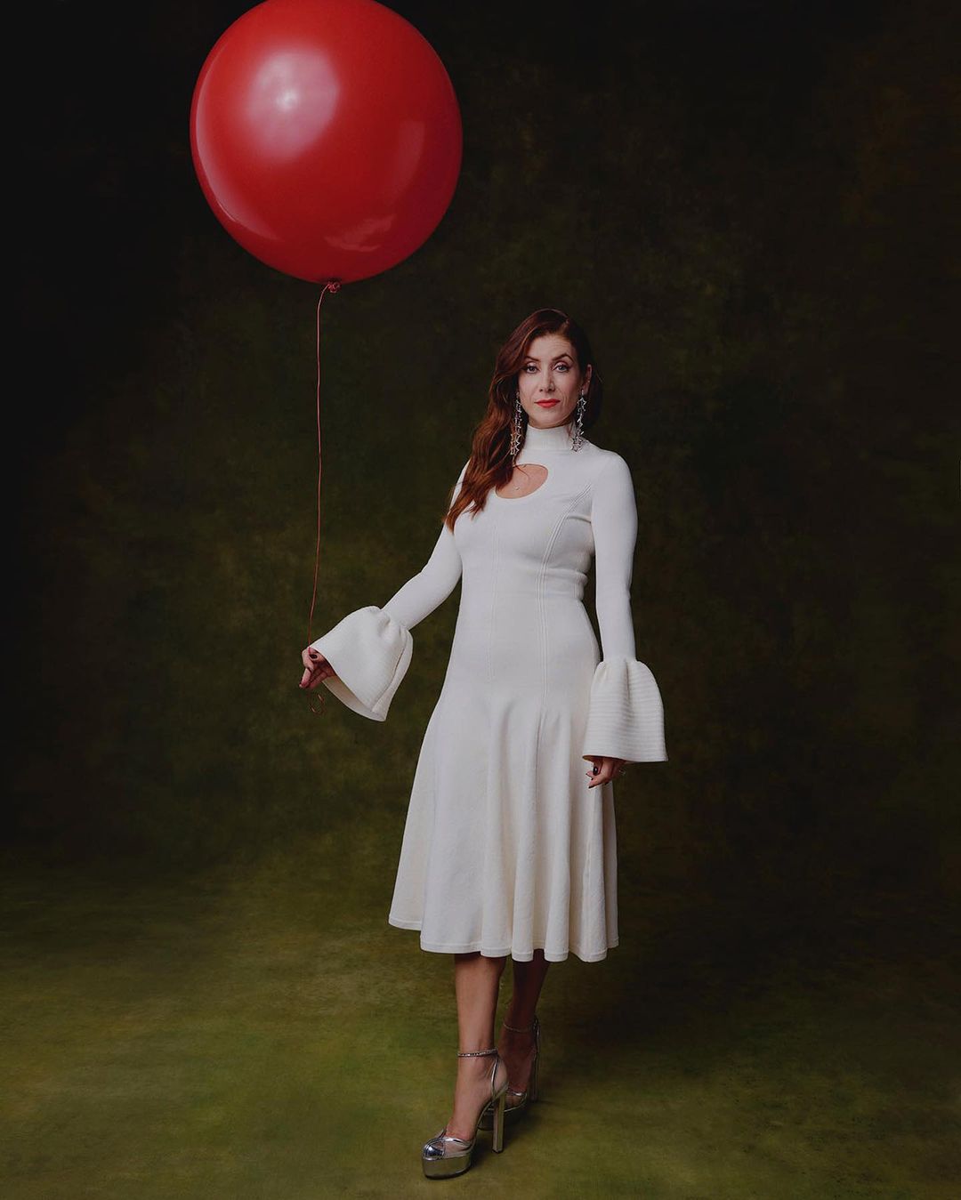 Woman in white dress poses with red balloon against textured background. Fashion shot with elegant dress and platform heels.