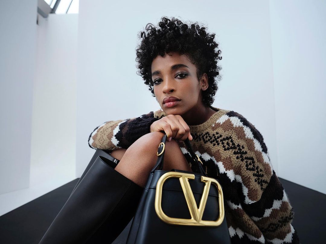 Adult female with curly dark hair poses with a black leather 'V' logo purse while wearing a patterned knit sweater and black leather boots.