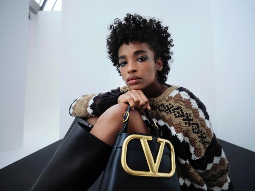 Adult female with curly dark hair poses with a black leather 'V' logo purse while wearing a patterned knit sweater and black leather boots.